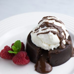Chocolate lava cake on a white plate with ice cream and chocolate drizzled on top. 3 raspberries on the side