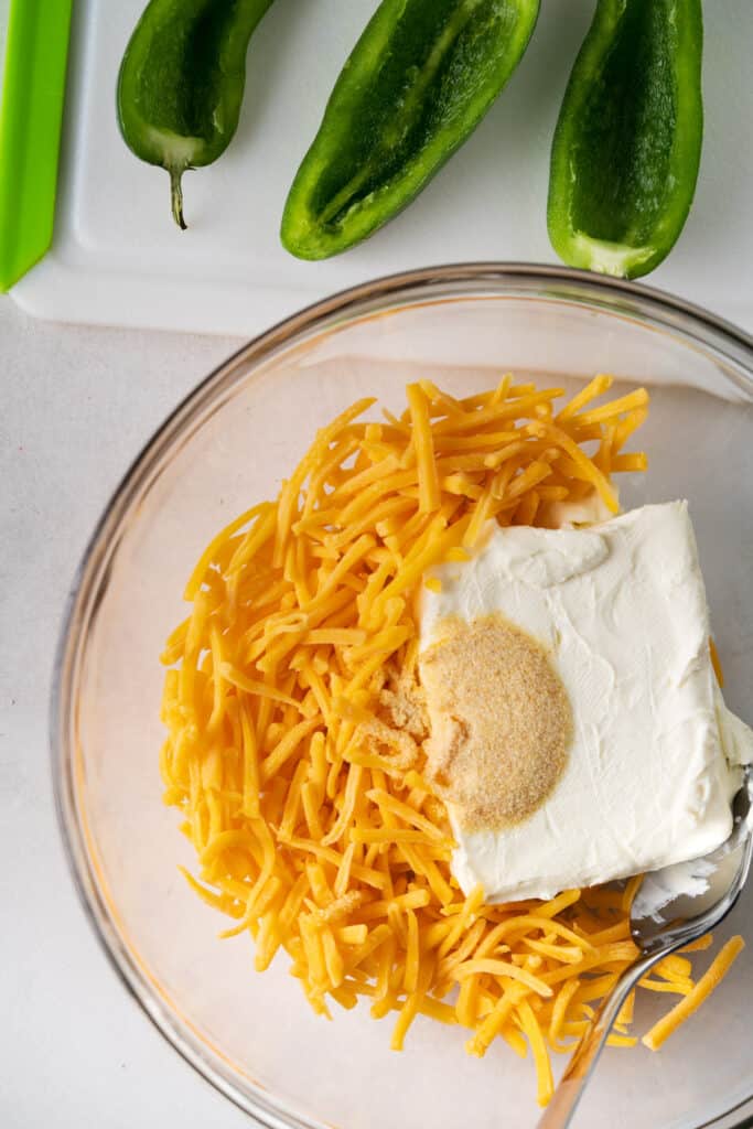 a clear bowl filled with shredded cheddar cheese, a block of cream cheese, and garlic powder