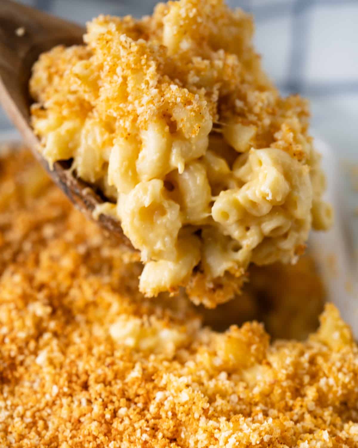 wooden spoon taking a spoonful of mac and cheese topped with breadcrumb from a casserole dish