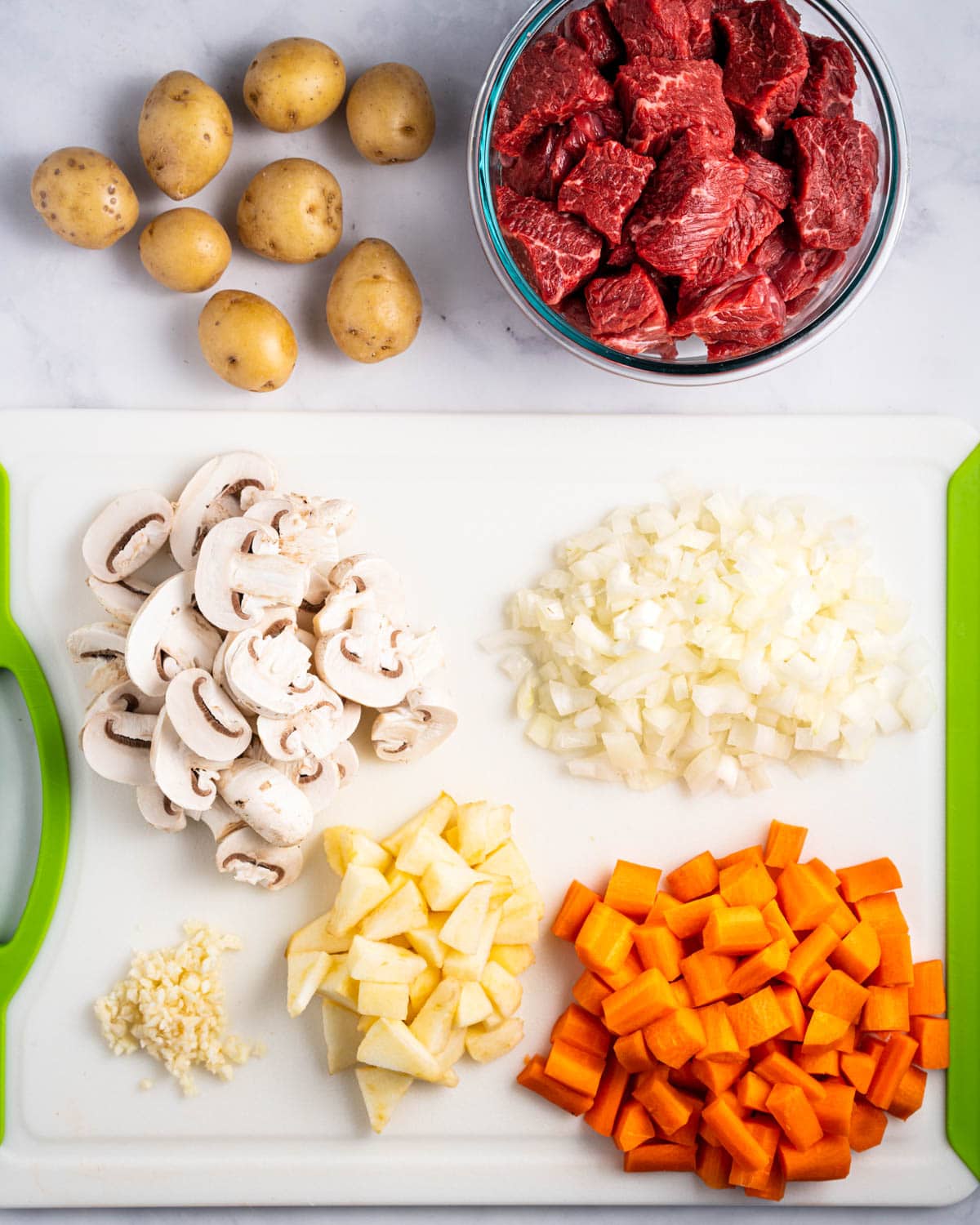 Cutting board with sliced mushrooms, diced onions, carrots, apples, and minced garlic. Bowl of beef cut into cubes.