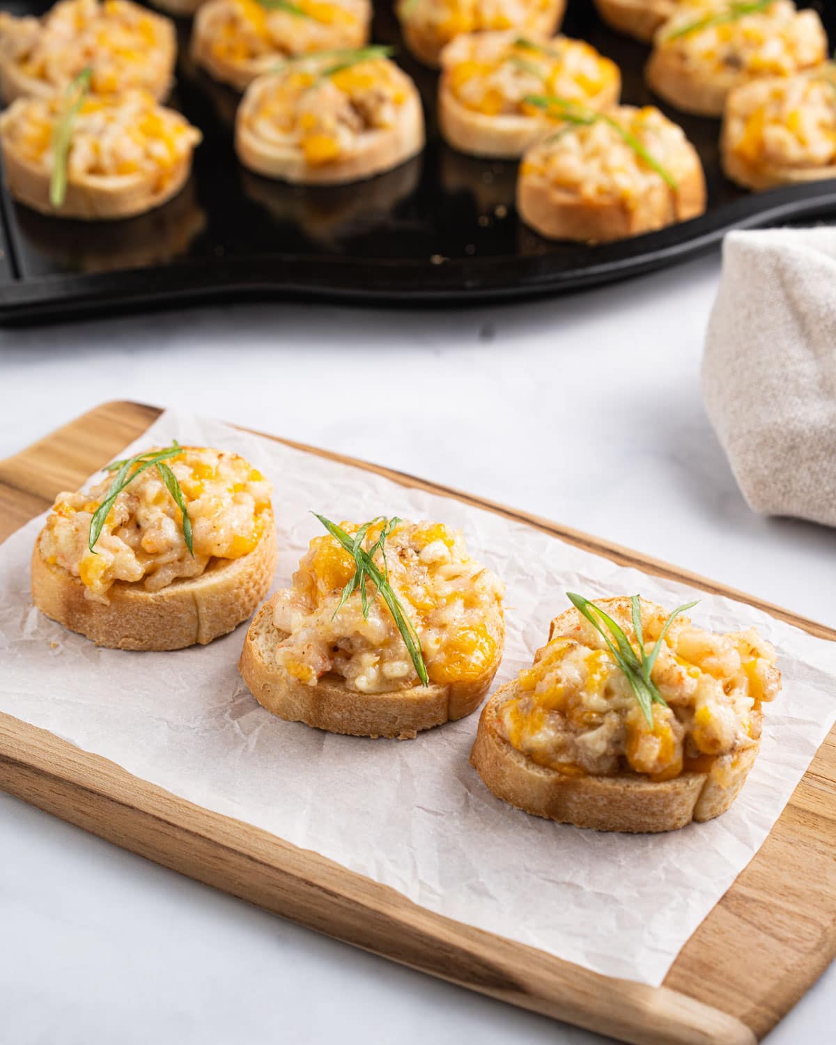 Tray of Vietnamese shrimp toasts in the background and 3 shrimp toasts on a serving board.