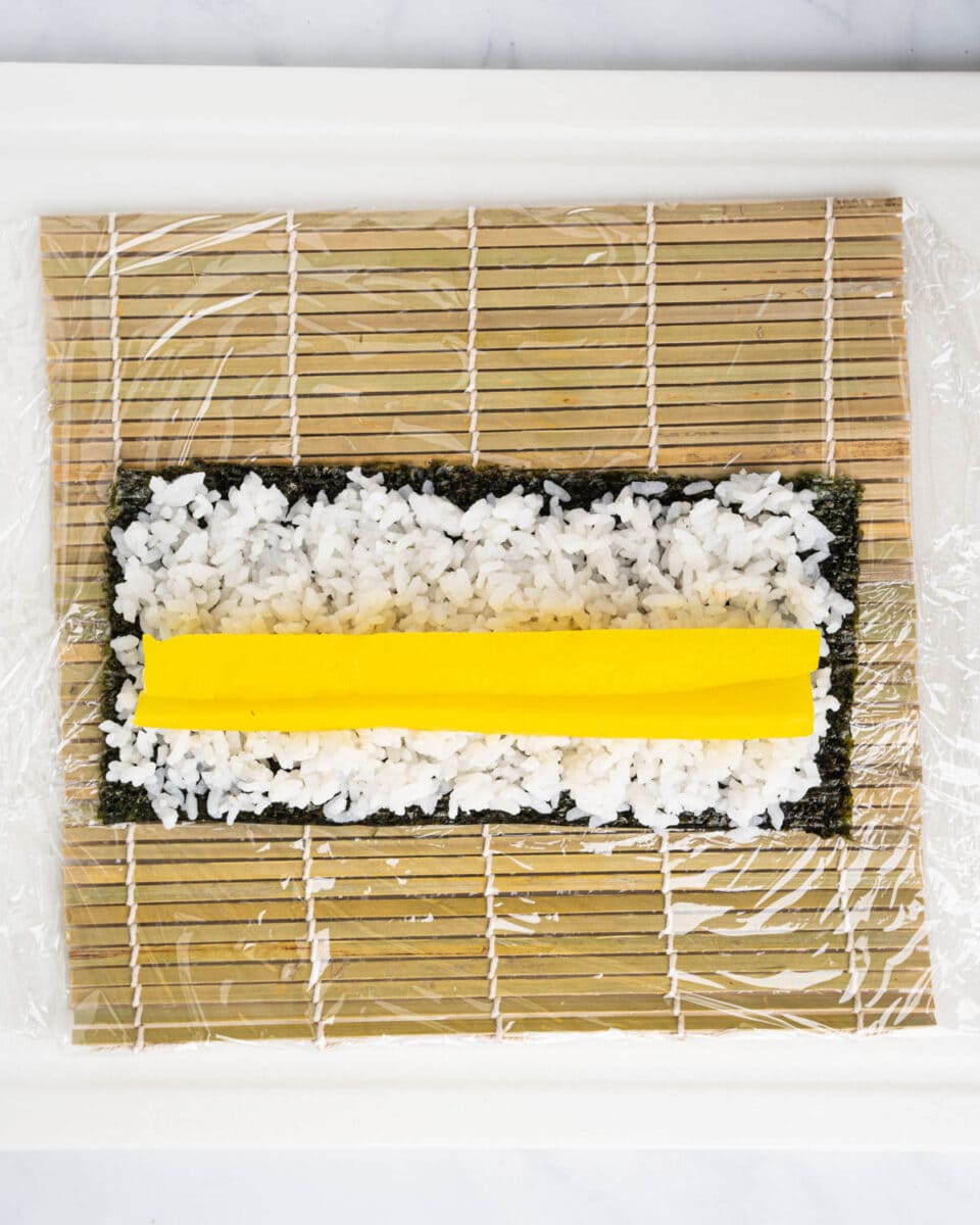 sushi mat with nori sheet topped with a layer of rice and Takuan in the middle.