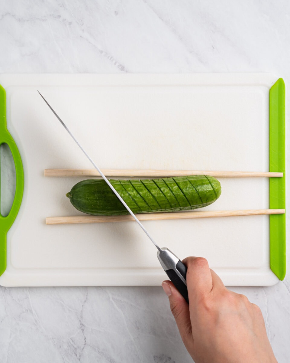 cucumber on a cutting board between two chopsticks and knife cutting diagonal 45 degree slits.