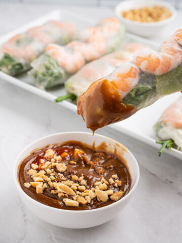 Dipping a spring roll into a bowl of peanut sauce topped with crushed peanuts and sriracha.