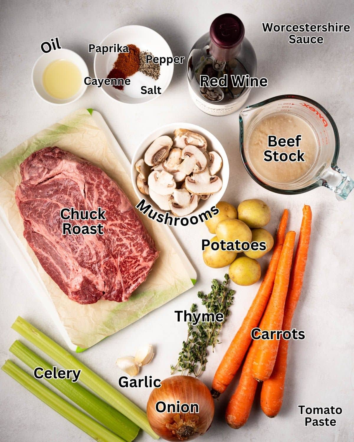 Ingredients needed for instant pot beef stew laid out on a white background: beef chuck roast, onions, garlic, celery, carrots, potatoes, mushrooms, beef stock, red wine, oil, Worcestershire sauce, tomato paste, salt, pepper, paprika, and ground cayenne.