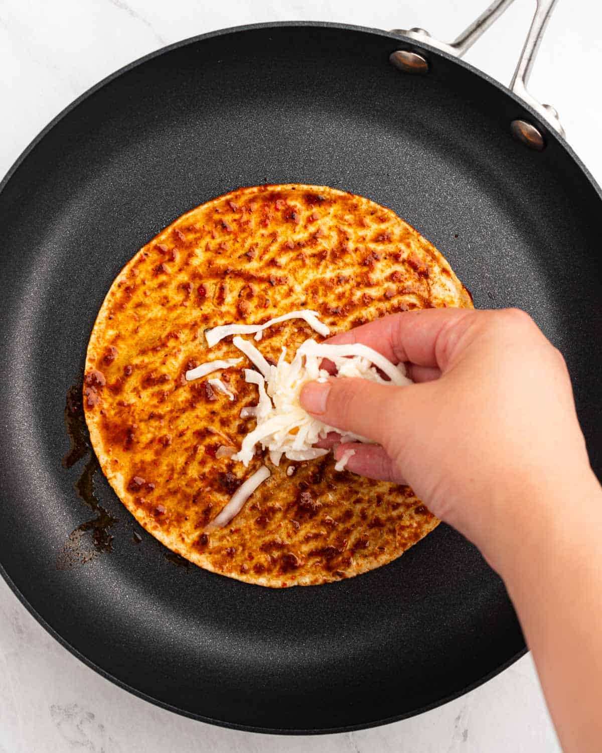 Hand spreading cheese on top of corn tortilla on a pan.