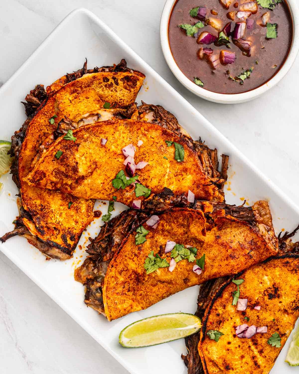 top down of birria tacos on a plate with side of lime and dipping sauce.