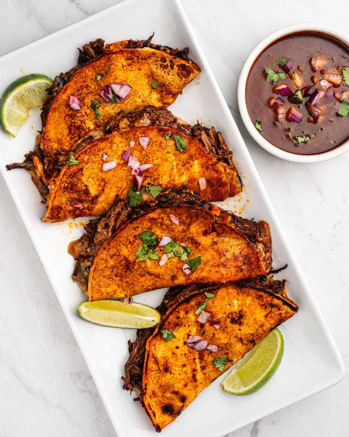 Four birria tacos on a plate with lime wedges and consommé dipping sauce.